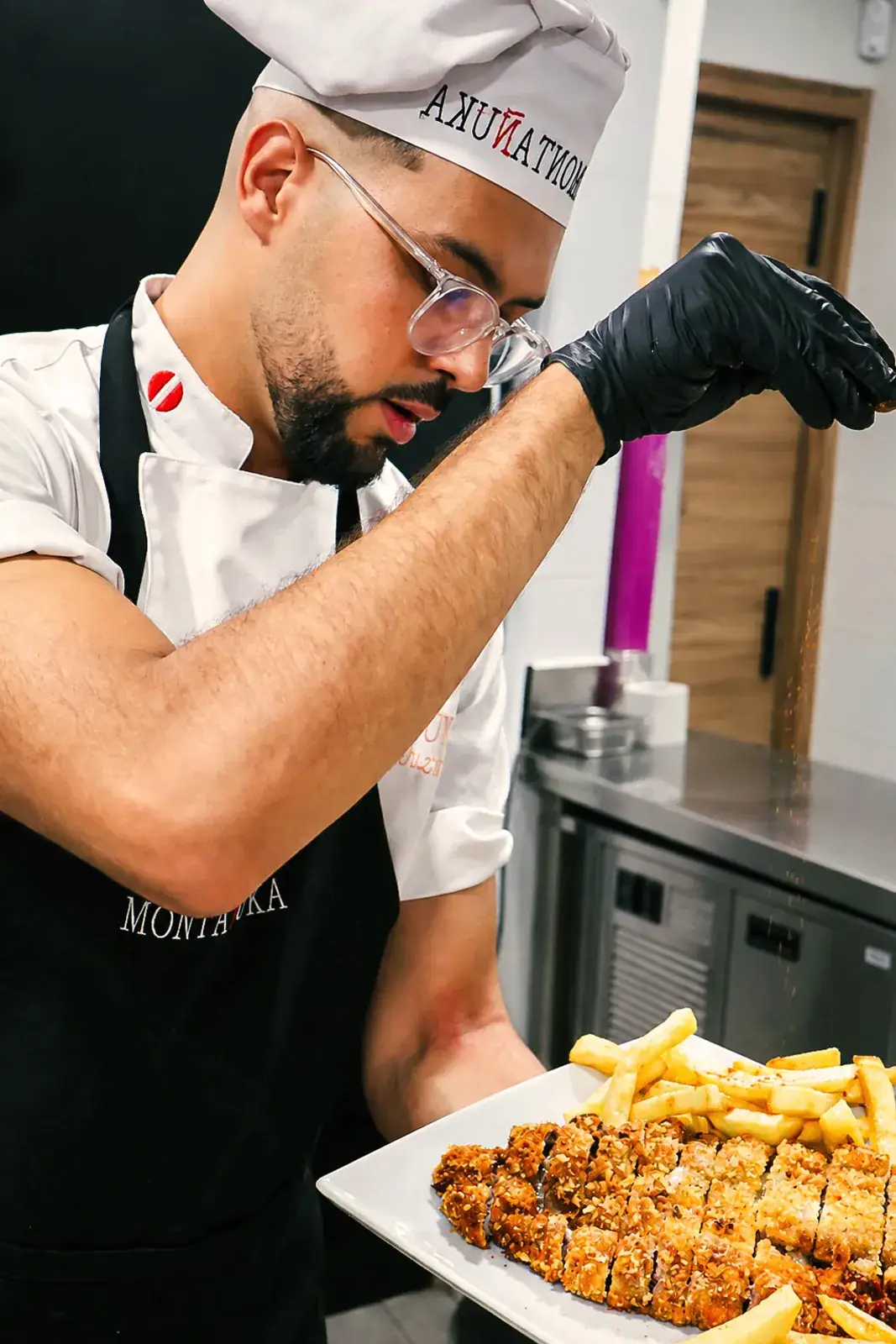 Chef de Montañuka con gorro y delantal negro espolvoreando especias sobre un cachopo cortado en porciones con patatas fritas
